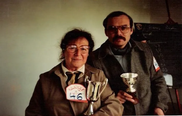 An older woman in glasses, wearing show number 46 and holding a trophy, next to a man with a moustache holding a smaller cup.