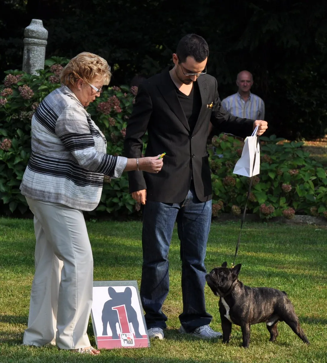 On a lawn, an older woman looks at a brindle French Bulldog while a man in a black jacket stands it next to the number 1 sign.